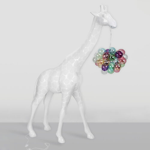 Glossy white giraffe sculpture with faint giraffe pattern holding a cluster of colorful translucent balloons in its mouth, standing on a plain gray surface against a light gray background