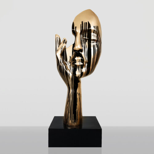 Abstract golden sculpture of a face with a hand touching the cheek, featuring black vertical streaks, mounted on a black base against a plain gray background
