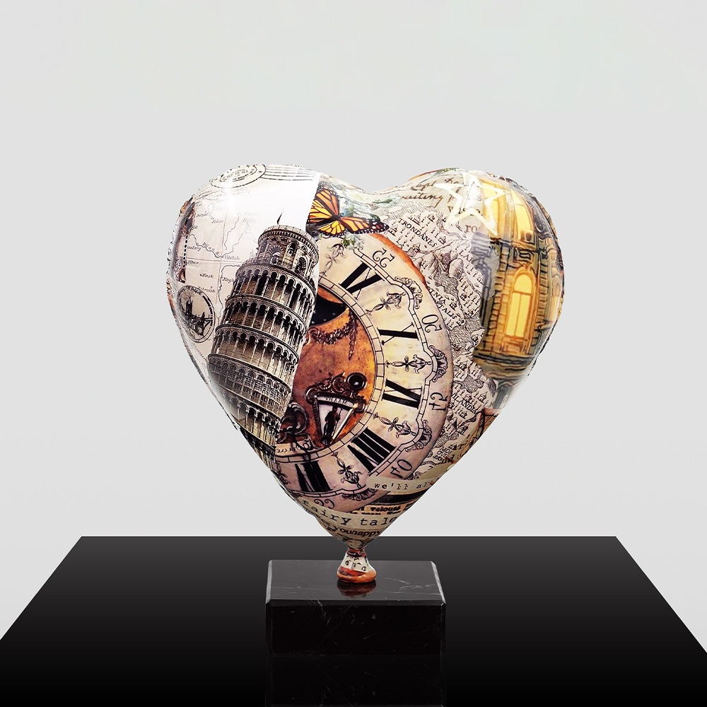 Decorative Heart-Shaped Balloon Figurine 3 Heart-shaped sculpture featuring collage designs including the Leaning Tower of Pisa, a vintage clock with Roman numerals, a butterfly, and an illuminated building, mounted on a black pedestal with a plain light gray background.