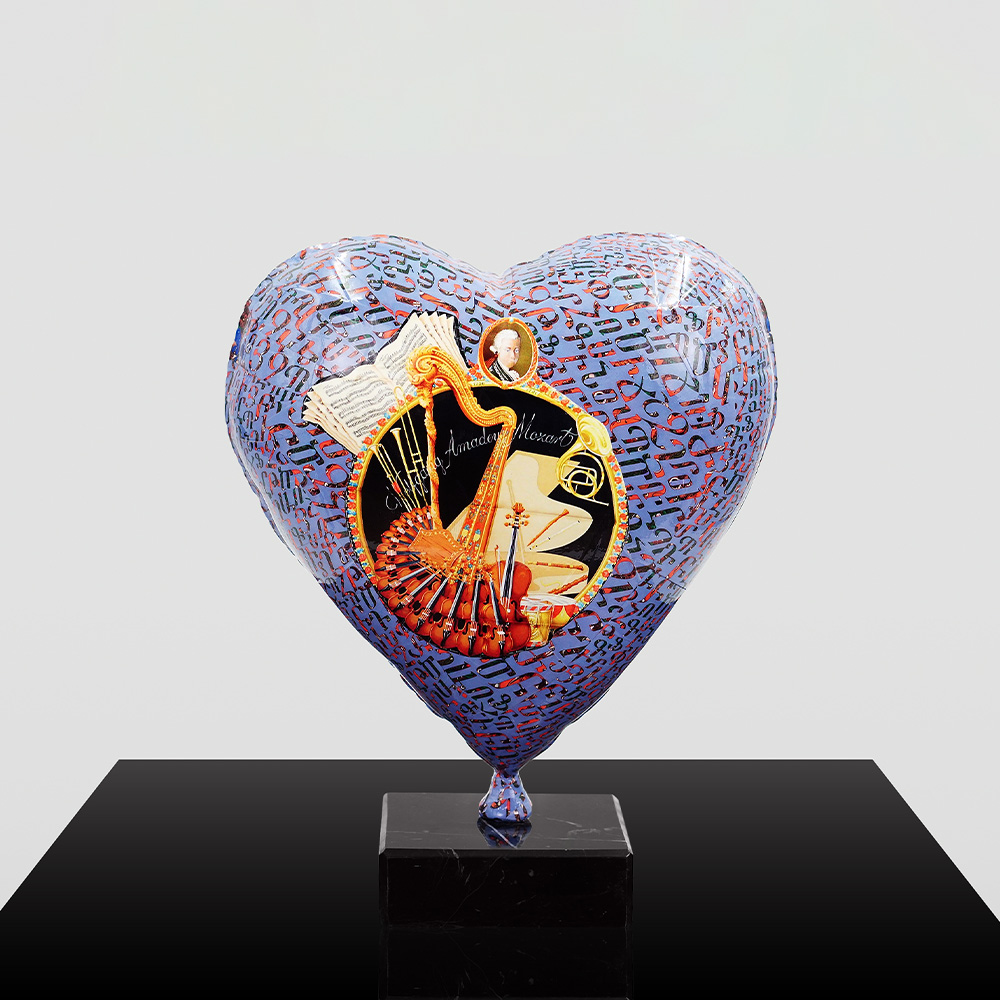 Decorative Heart-Shaped Balloon Figurine 5 Heart-shaped balloon sculpture decorated with musical notes and instruments, including violins and a harp, with a small portrait at the top and the name 'Amadeus Mozart' in the center, mounted on a black marble base against a plain background.