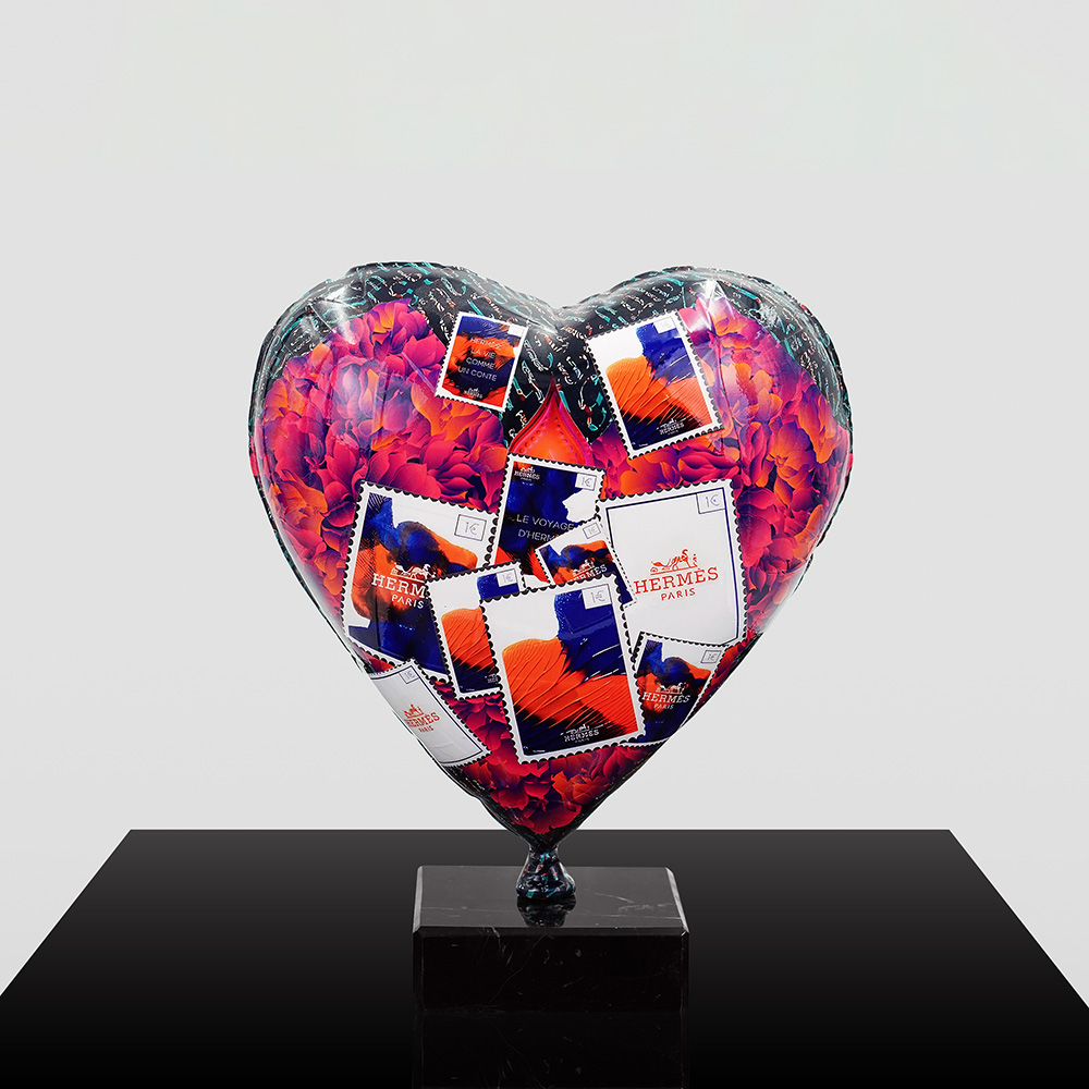 Decorative Heart-Shaped Balloon Figurine 7 A heart-shaped sculpture decorated with colorful flower patterns and Hermes Paris postage stamps, mounted on a black marble base against a plain light gray background.