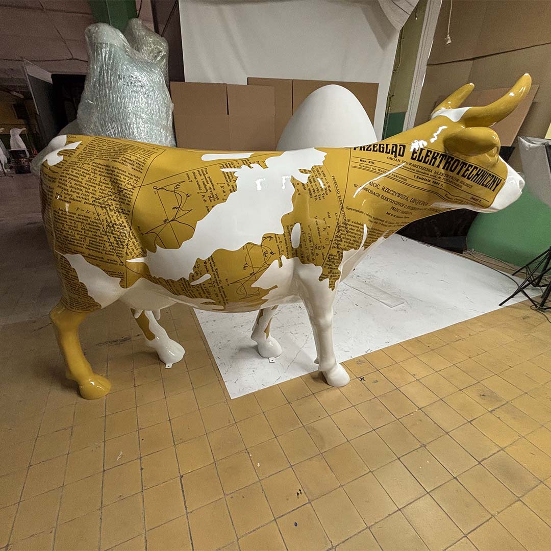 Figura krowy stojąca na żółto-brązowych kafelkach, pokryta białymi i żółtymi wzorami przypominającymi strony tekstu z polskimi napisami.