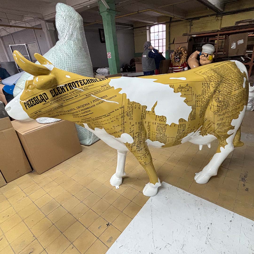 rzeźba krowy ozdobiona wzorami przypominającymi stare gazety i teksty techniczne, stojąca w pomieszczeniu magazynowym z osobami i dużymi figurami w tle