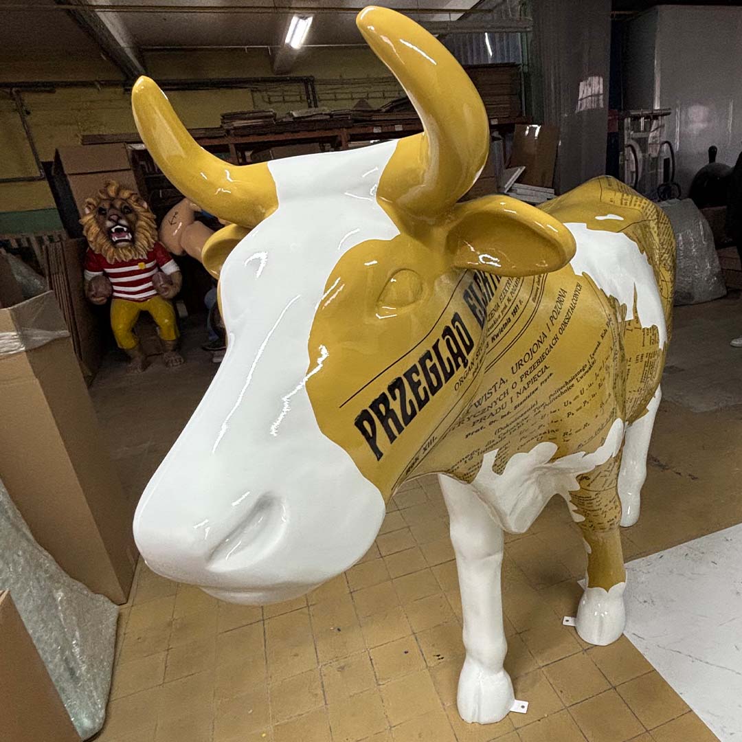 Glossy cow sculpture painted white with yellow patches featuring vintage newspaper print, standing indoors on a tiled floor with shelves and lion and human-like statues in the background.