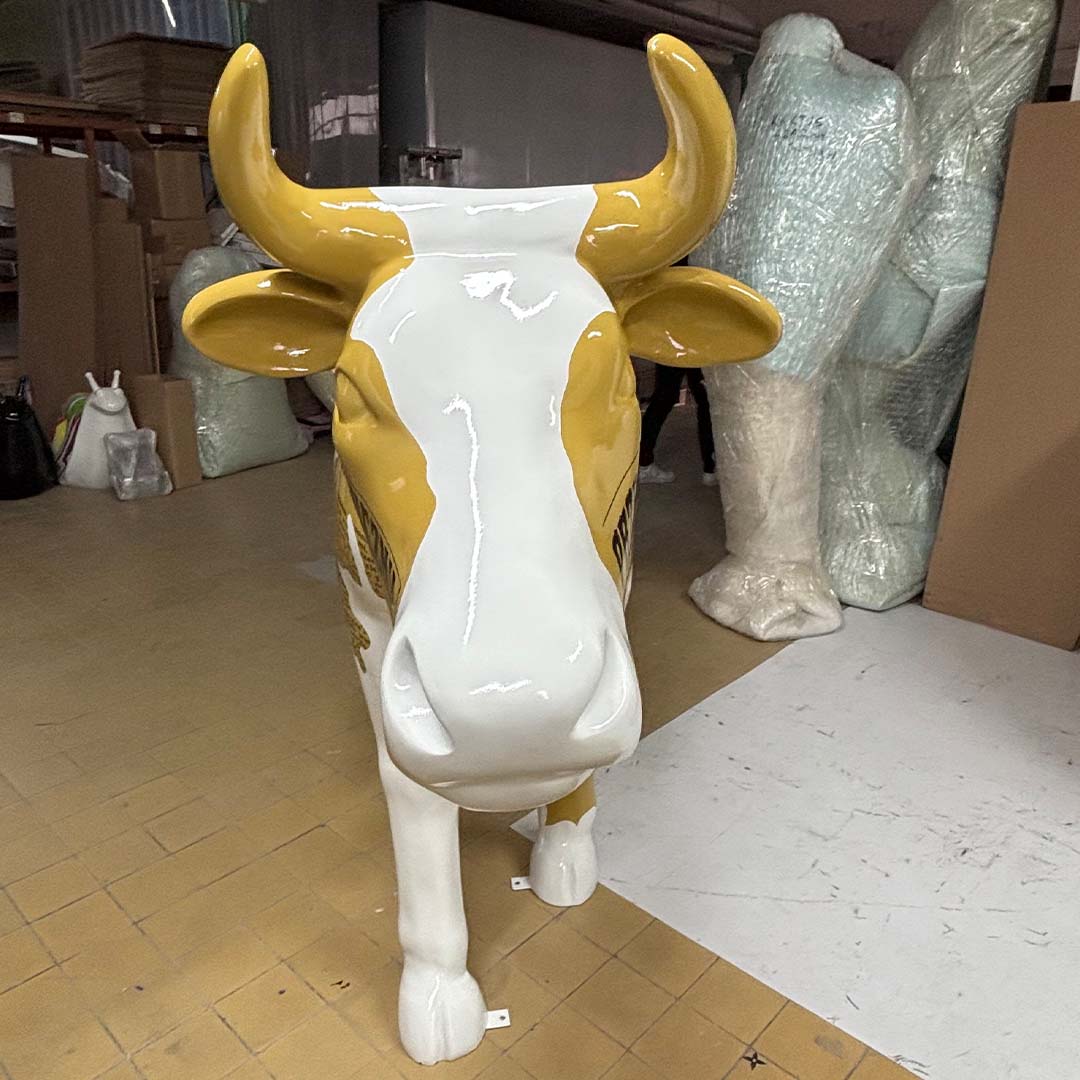 Figura krowy stojąca na podłodze z żółto-białym malowaniem, w magazynie z owiniętymi w folię przedmiotami w tle