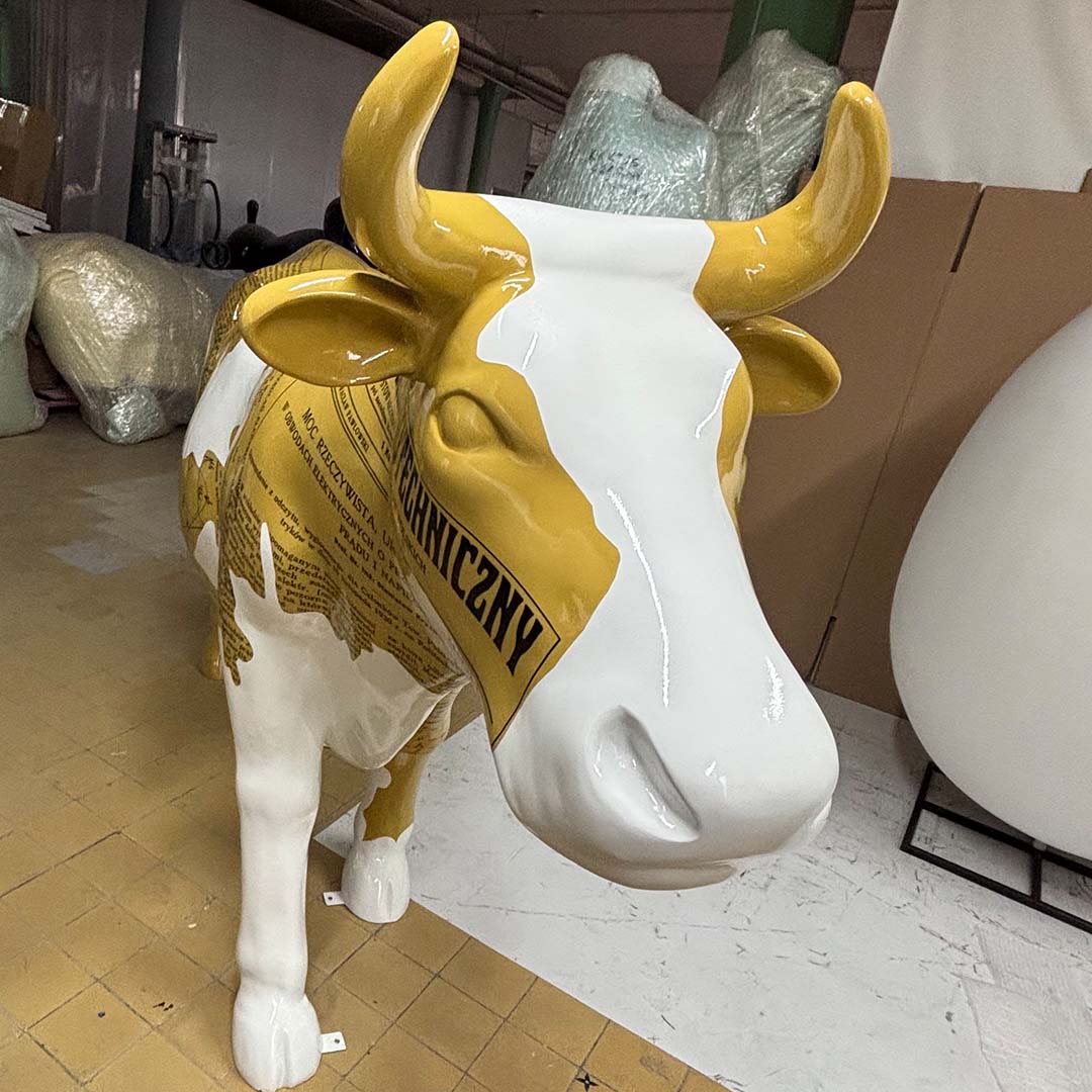 Rzeźba krowy w kolorze białym i żółtym z nadrukiem tekstu, stojąca na podłodze w pomieszczeniu magazynowym lub warsztatowym, w tle kartony i owinięte folie ochronne