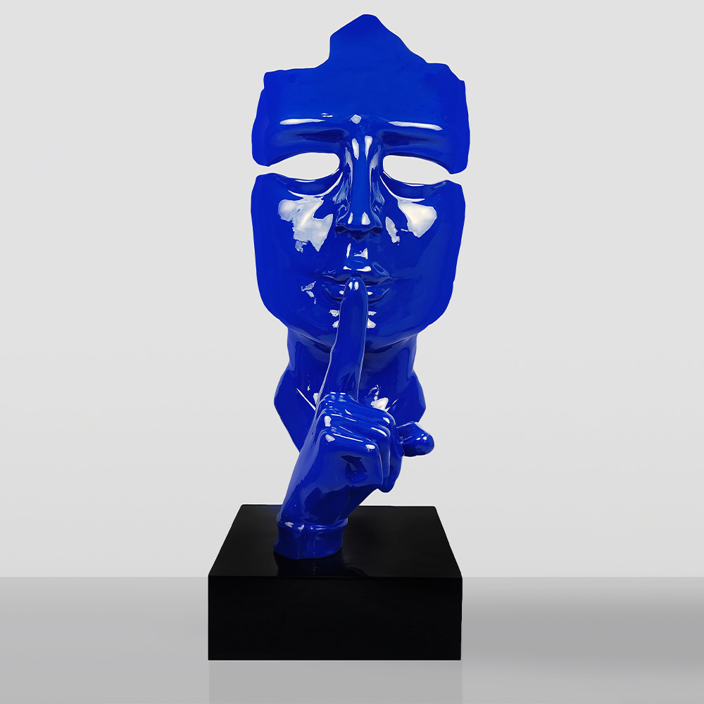 Large Mask on Stand – Blue 1 Glossy blue sculpture of a face with closed eyes and a raised finger held to the lips in a shushing gesture, mounted on a black base against a light gray background