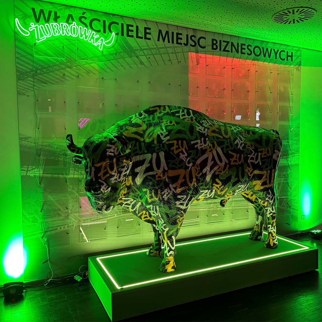 A large bull sculpture covered in colorful graffiti-style text with the word ZU repeatedly written, displayed on a lit green pedestal in a room with green lighting and a sign above reading WŁAŚCICIELE MIEJSC BIZNESOWYCH.