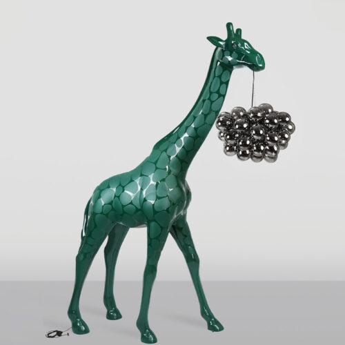 Green giraffe sculpture with a pattern of darker green spots holding a cluster of metallic silver spheres hanging from its mouth by a thin string against a plain gray background.